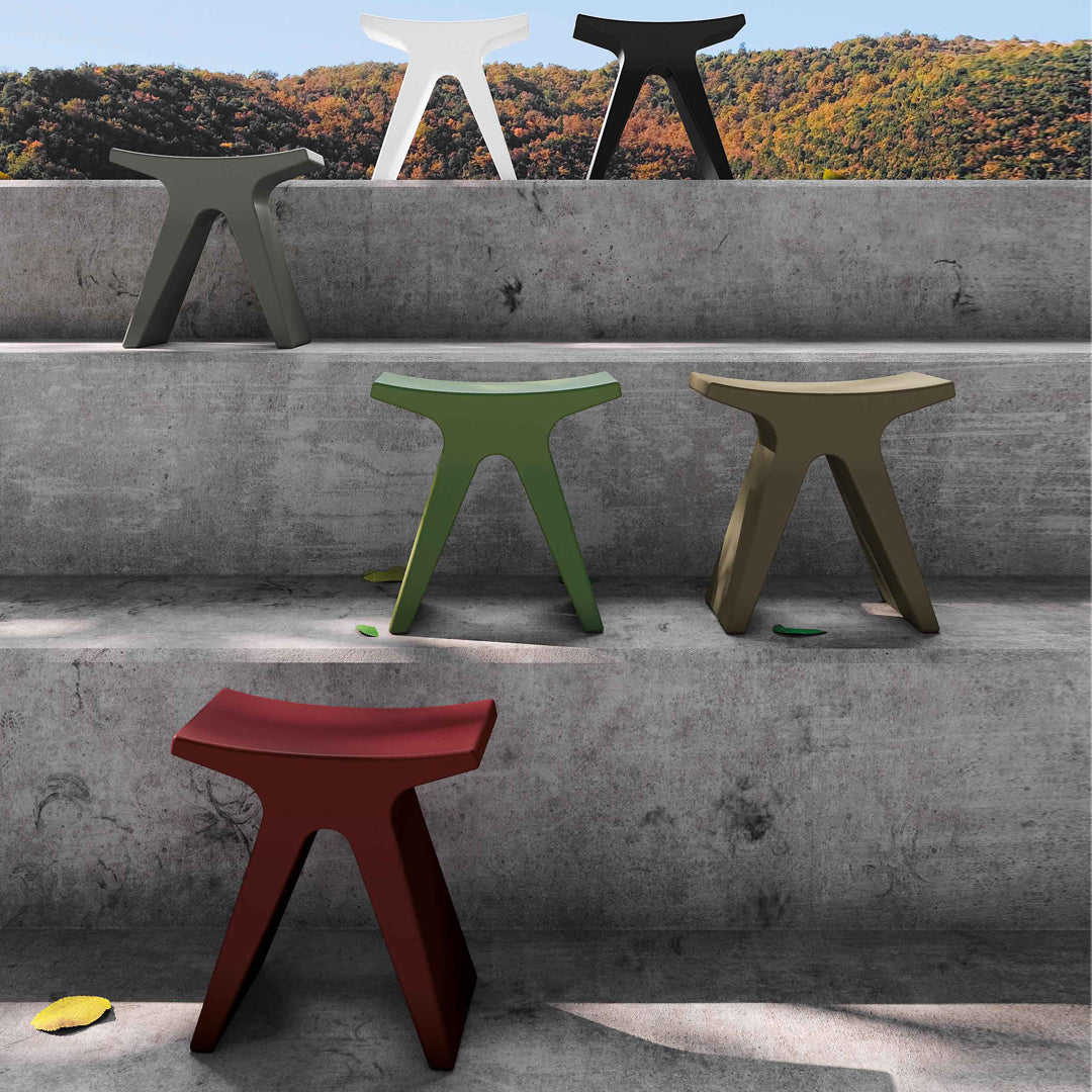 Colorful stools in red, green, olive, white, and gray with modern, smooth shapes on a textured concrete surface.