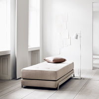 Light beige bed with a smooth fabric texture and a contrasting brown pillow against a minimalist white background.