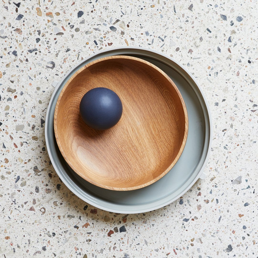 Wooden bowl with natural finish, nestled in a light gray circular dish, topped by a matte navy ball. Minimalist design.
