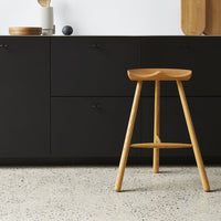 Natural wooden bar stool, light oak finish, complemented by a sleek black cabinet and textured light speckled flooring.