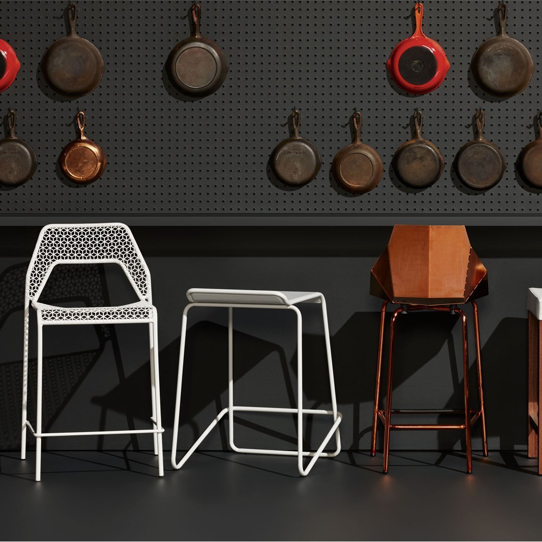"Three stylish barstools in white, copper, and wood against a black pegboard backdrop with hanging cookware."