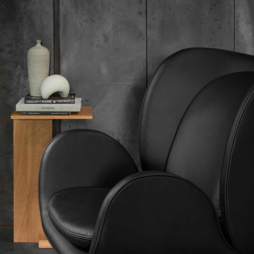 Modern black leather chair with smooth curves, against a textured gray wall and warm wooden side table.