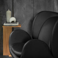 Modern black leather chair with smooth curves, against a textured gray wall and warm wooden side table.