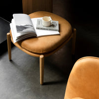 Tan upholstered stool with rounded edges, set against a dark floor, featuring subtle textures and clean design.
