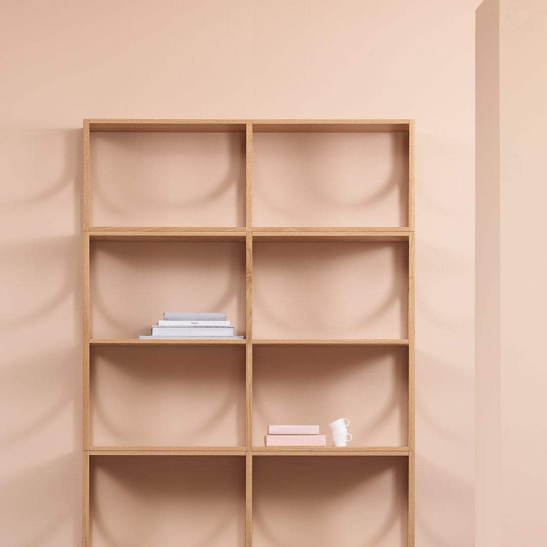 Light wood bookshelf against a soft peach wall, displaying white and pastel book colors in a minimalistic pattern.