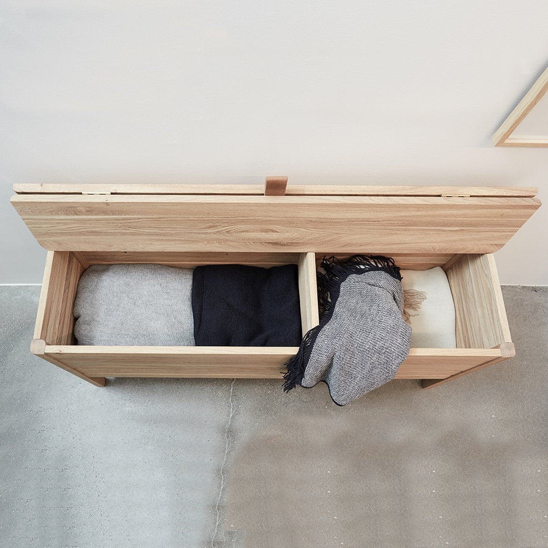 Light wood storage bench with grey, black, and white clothing items visible inside. Modern, minimalist design.