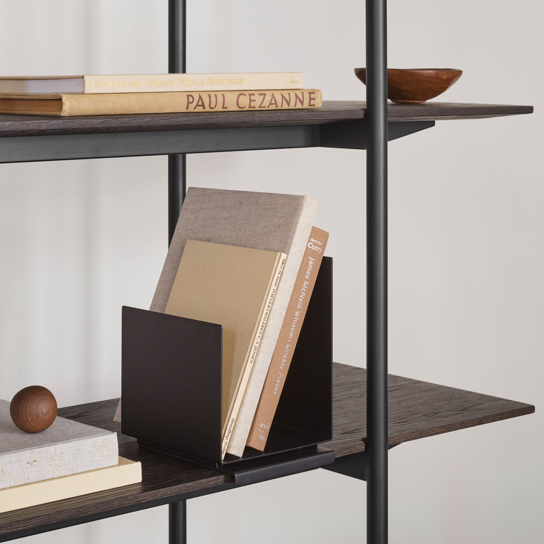 Modern black metal bookends with beige and gray books, set against a dark wood shelf. Decorative bowl in warm tones above.