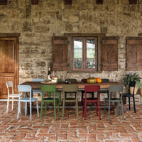 Colorful dining chairs in green, red, gray, and white against a rustic stone wall and terracotta floor.