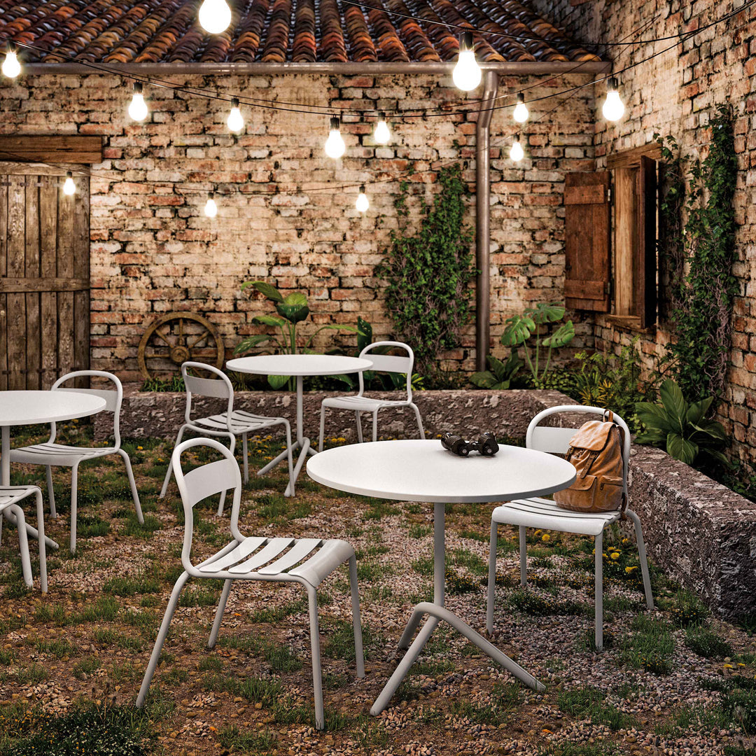 "White metal outdoor tables and chairs against a rustic brick wall, illuminated by warm string lights and greenery."