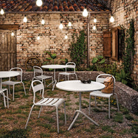 "White metal outdoor tables and chairs against a rustic brick wall, illuminated by warm string lights and greenery."