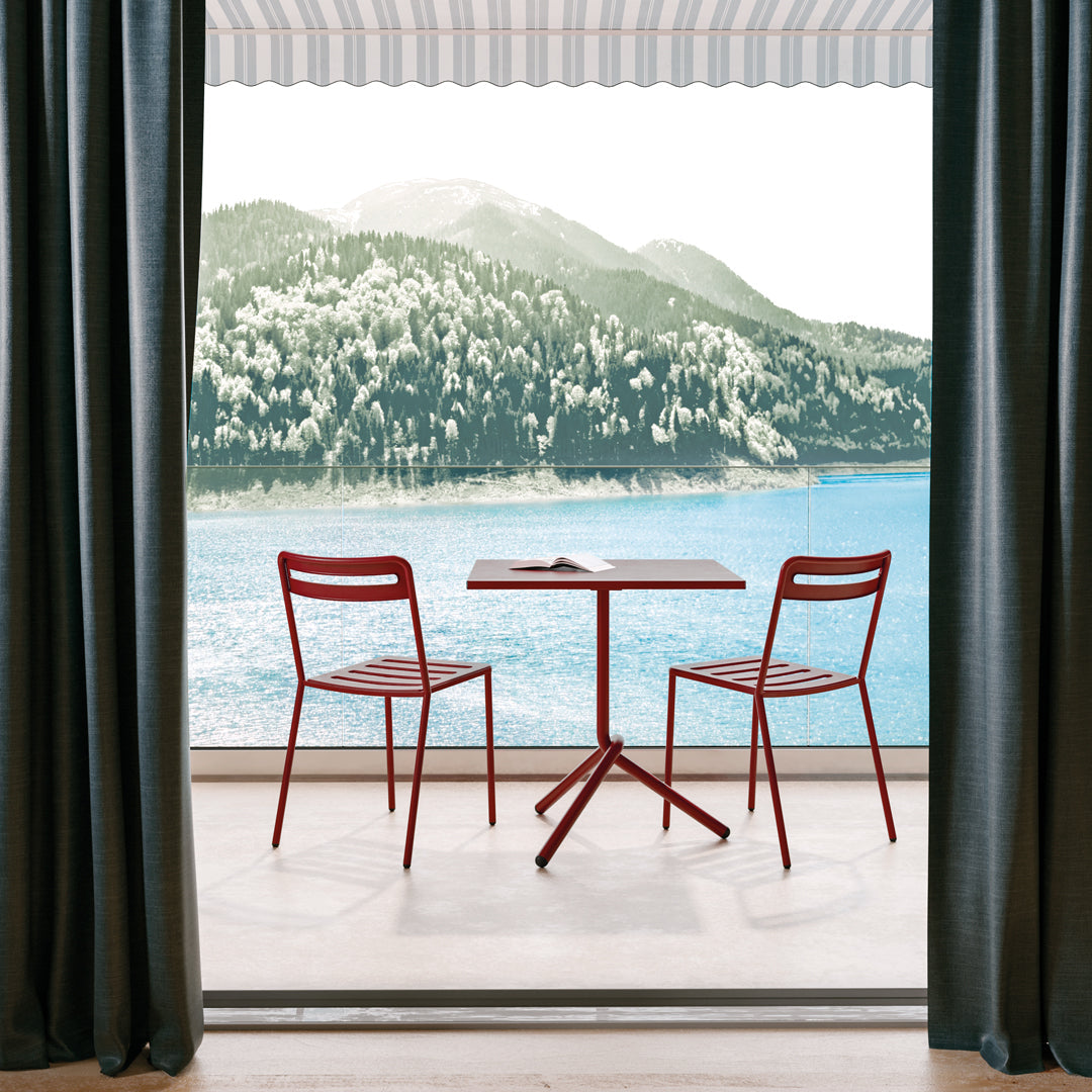 Red metal chairs at a contemporary table, set against a backdrop of green mountains and blue water, with striped curtains.