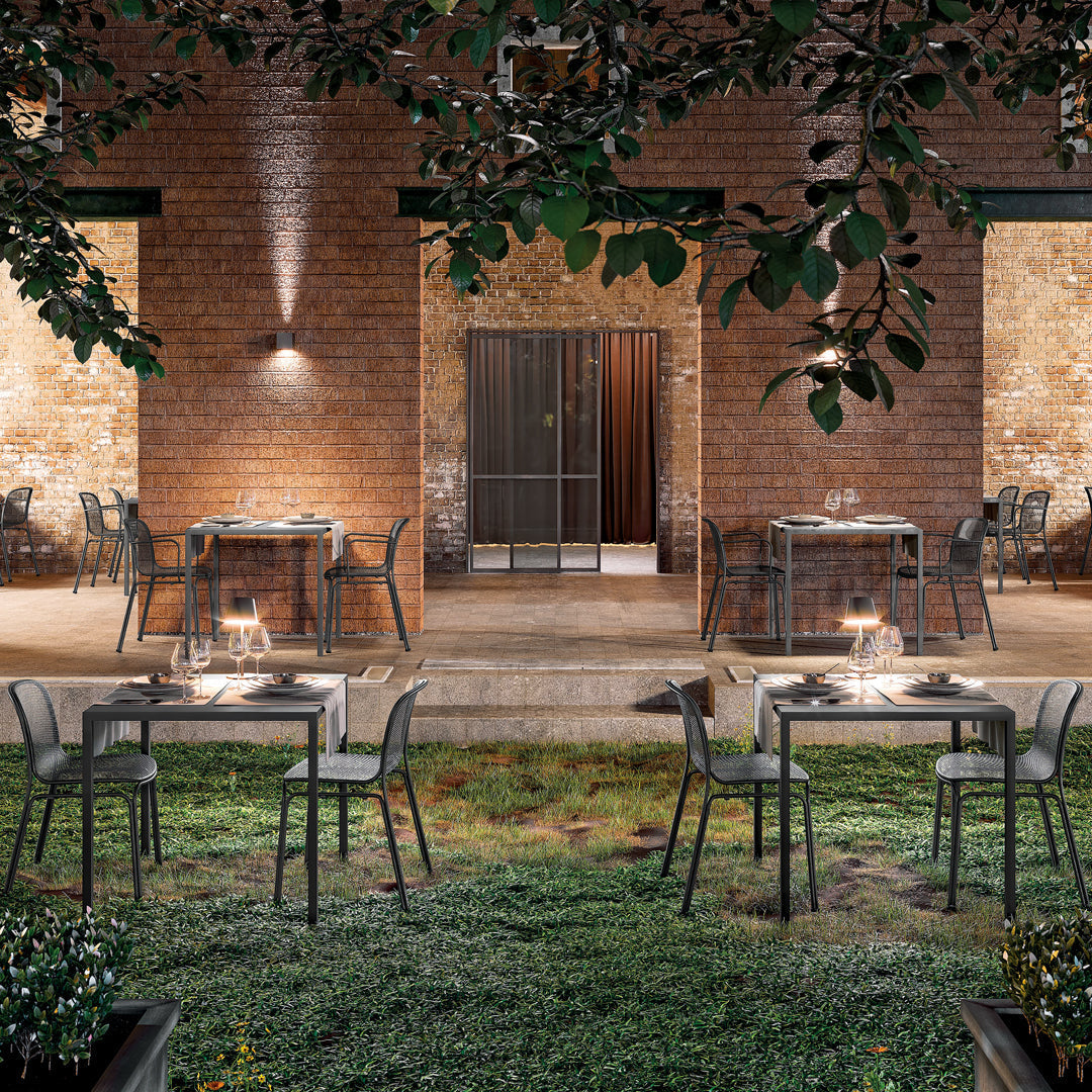 Alfresco dining set featuring sleek, dark metal chairs and glass tables against a rustic brick and green backdrop.