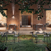 Alfresco dining set featuring sleek, dark metal chairs and glass tables against a rustic brick and green backdrop.