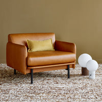 Brown leather armchair with a yellow accent pillow on a textured beige and brown rug, against a tan wall.