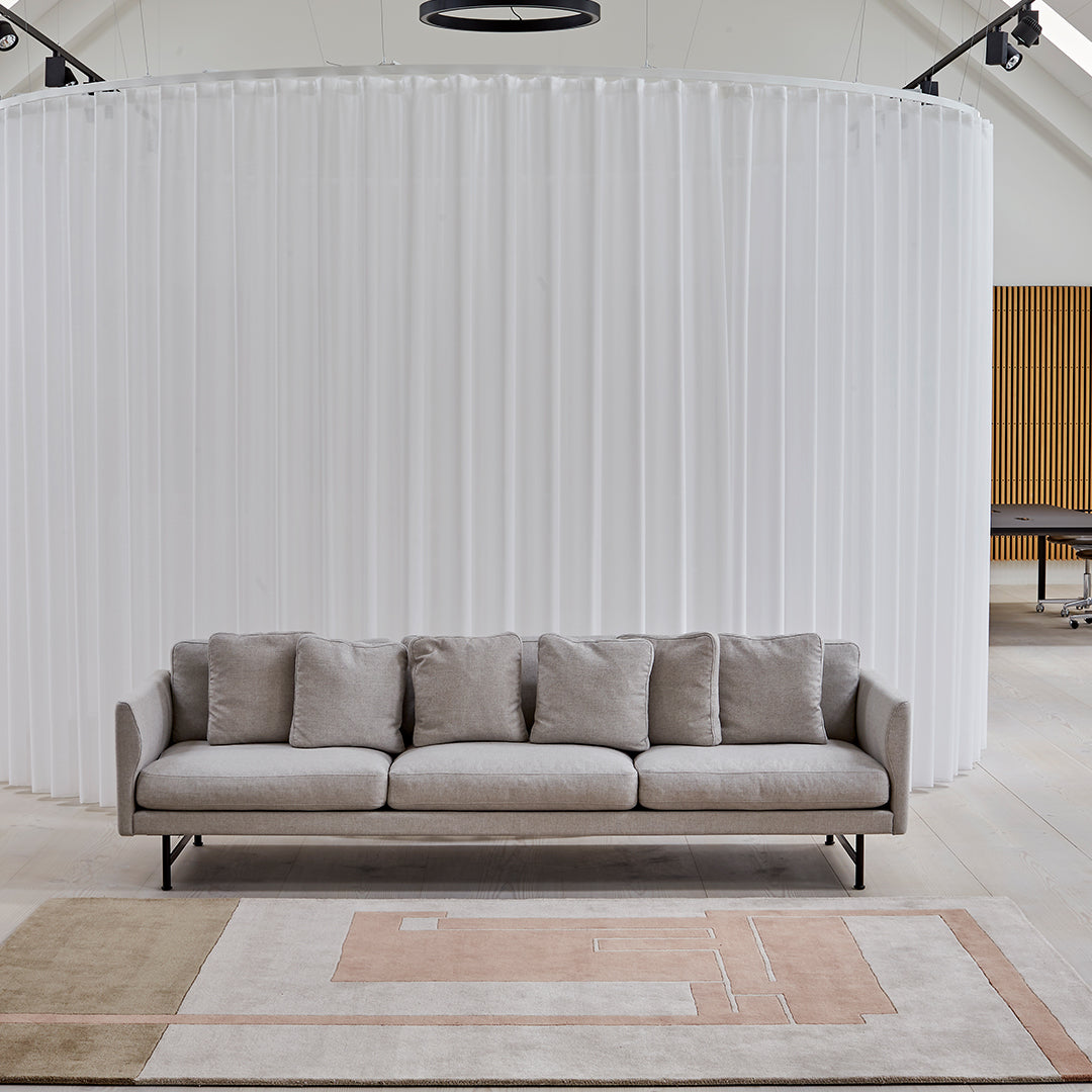 Light gray sofa with plush cushions, set against a backdrop of white curtains and a subtle patterned rug in beige and pink.
