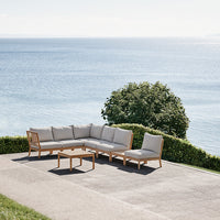 Gray upholstered outdoor sectional sofa with light wood frame against a serene coastal backdrop.