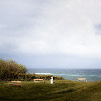 "Natural wood benches on grass, contrasting against a cloudy sky and tranquil ocean backdrop."