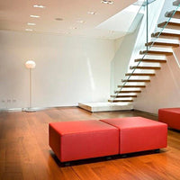 Red upholstered ottomans on warm wooden floor, with minimalist white walls and modern glass staircase.