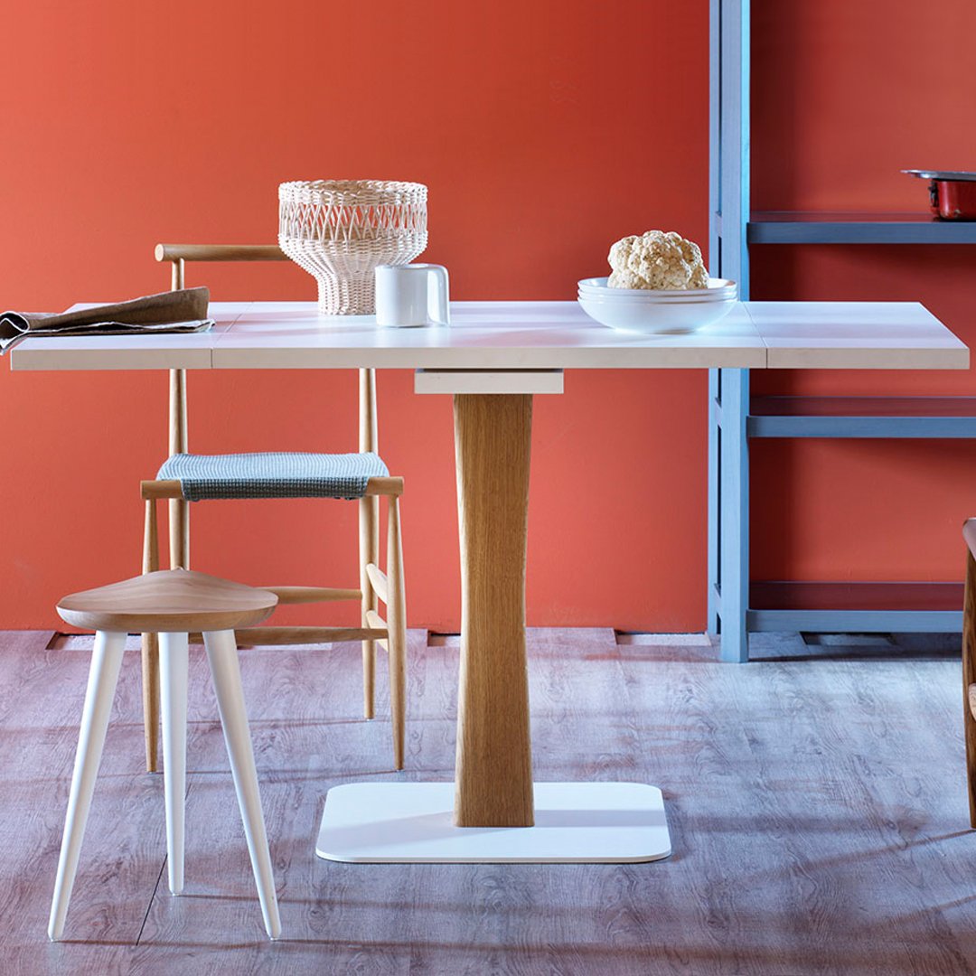 Modern white dining table with a natural wood pedestal, surrounded by colorful chairs against a warm orange wall.