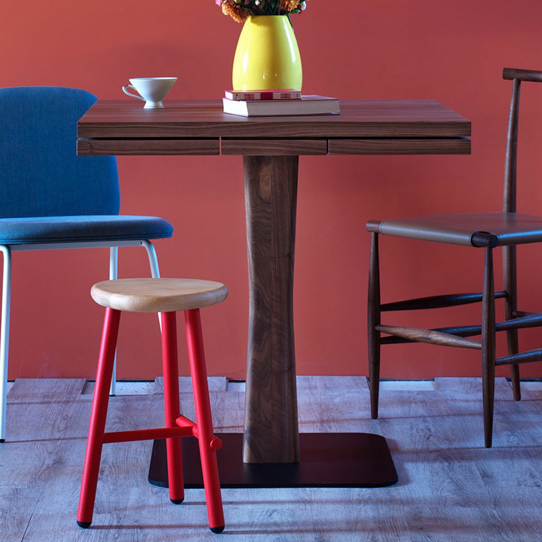 Modern wooden table with two-tone finish, paired with a red stool and blue chair against a vibrant orange backdrop.