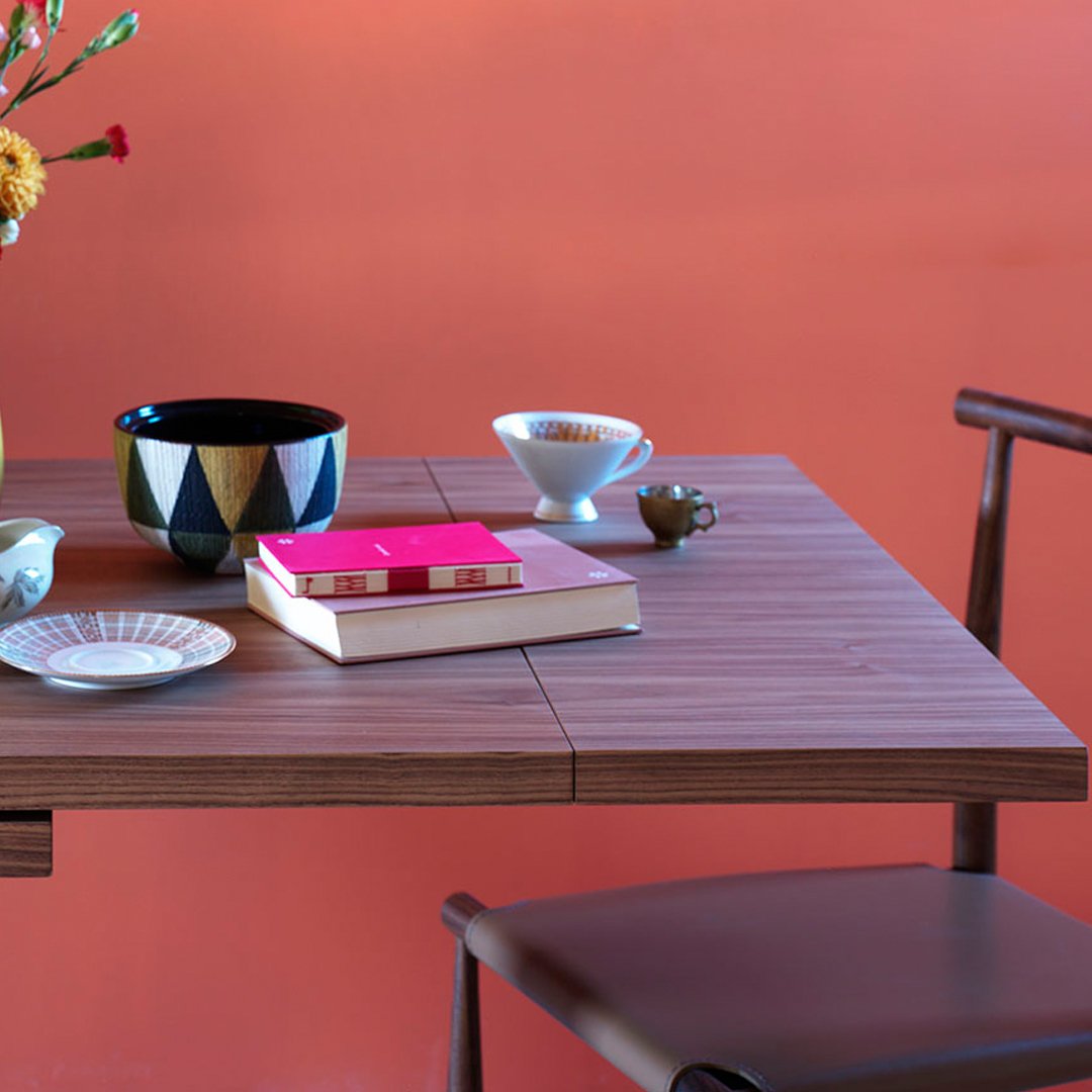 Wooden table with geometric patterned bowl, colorful books, and minimalistic dishware against a coral wall backdrop.