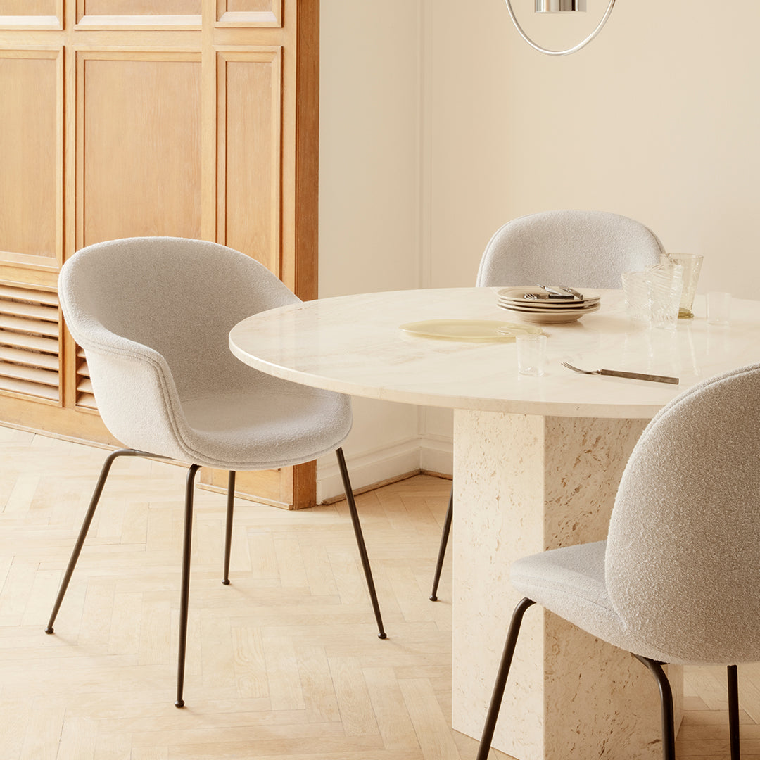 Light gray upholstered chairs around a round beige marble table with subtle textures, set in a warm-toned interior.