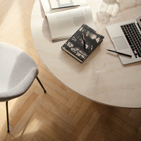 Round light-colored table with a patterned gray chair and scattered books on a warm wood floor. Elegant modern design.
