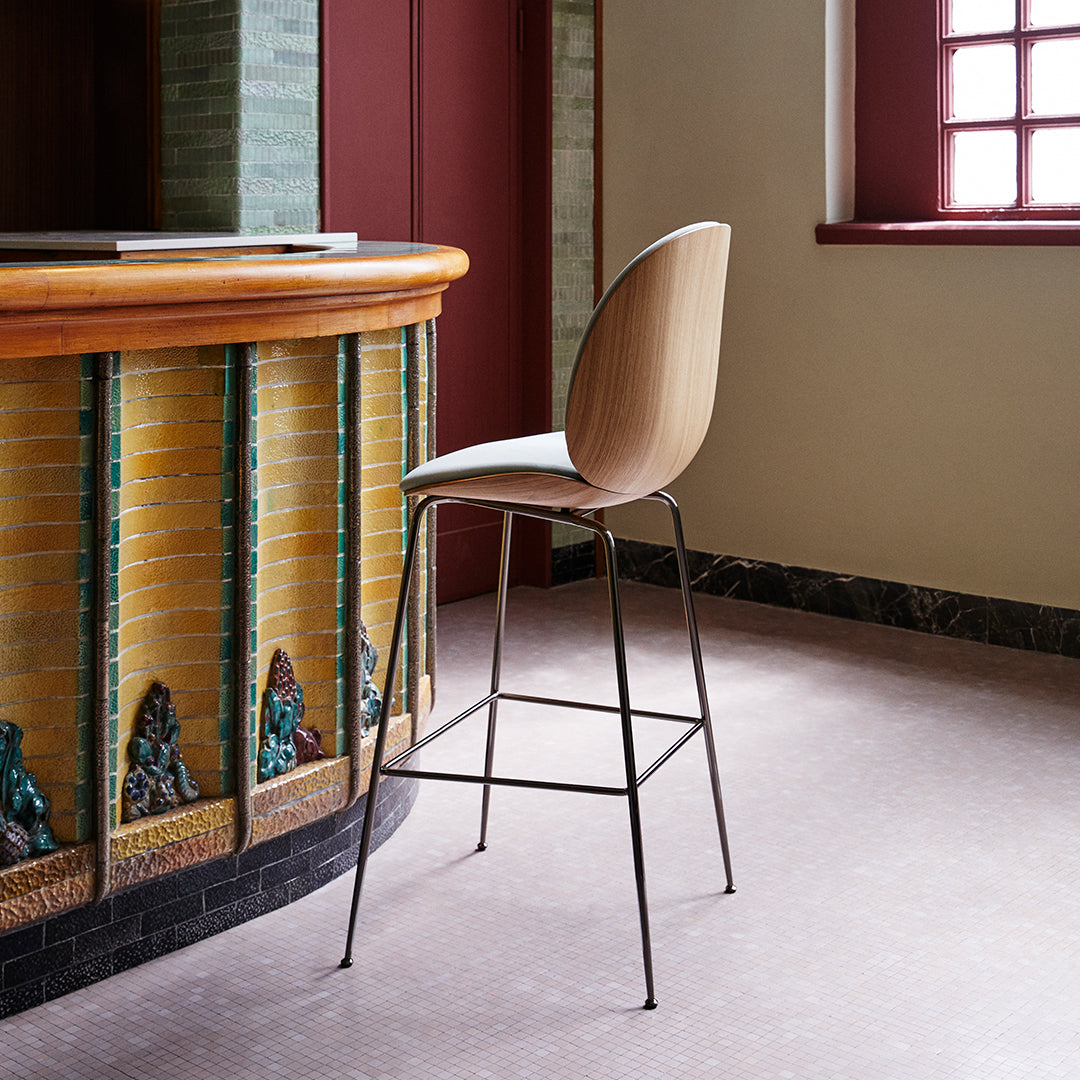 Stylish beige bar stool with sleek black legs, set against a colorful tiled bar backdrop. Modern and elegant design.