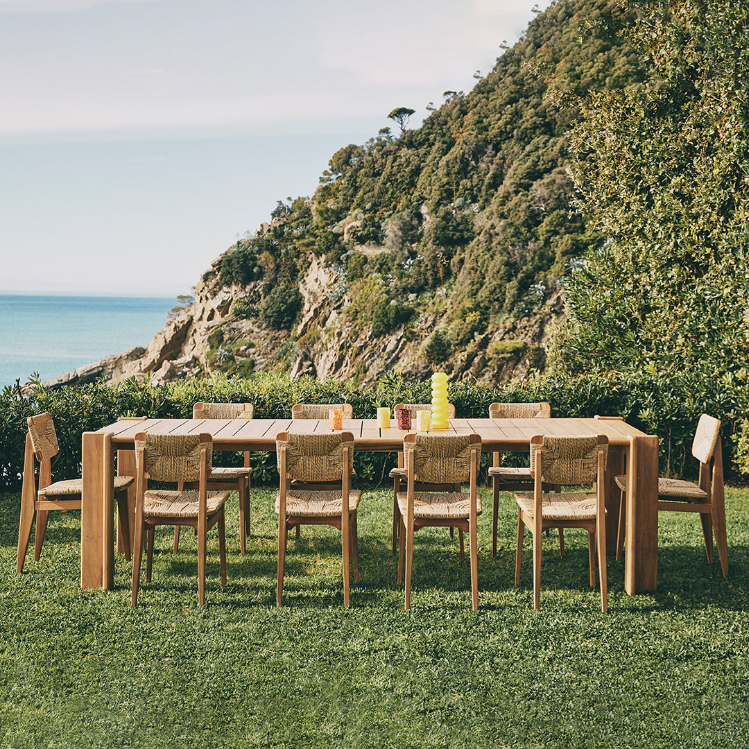 Wooden outdoor dining set with sandy beige cushions and a natural finish, surrounded by lush greenery and ocean views.