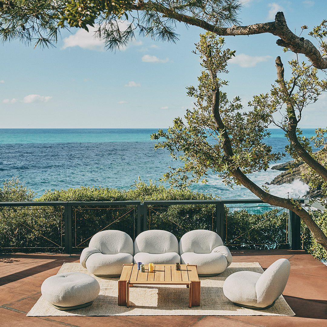 Soft white lounge chairs and poufs on a tan rug, set against a vibrant blue ocean and green foliage.