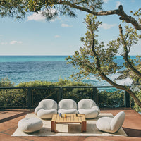 Soft white lounge chairs and poufs on a tan rug, set against a vibrant blue ocean and green foliage.