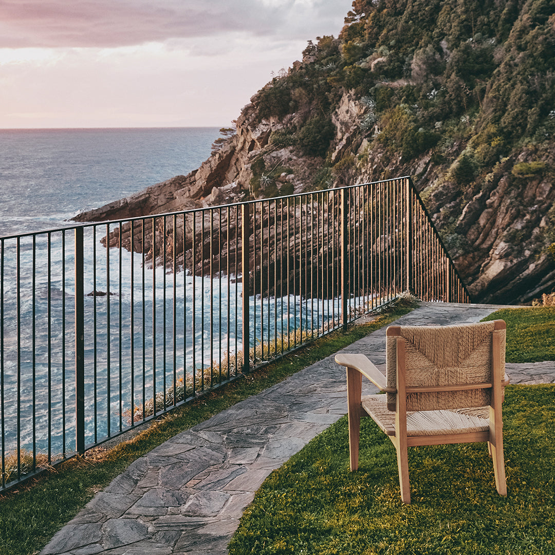 Wooden chair with natural finish, set against a backdrop of ocean waves and rocky cliffs at sunset.