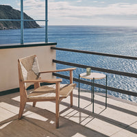 "Natural wood chair with woven detail on a balcony, complemented by a sleek circular side table against a blue ocean backdrop."