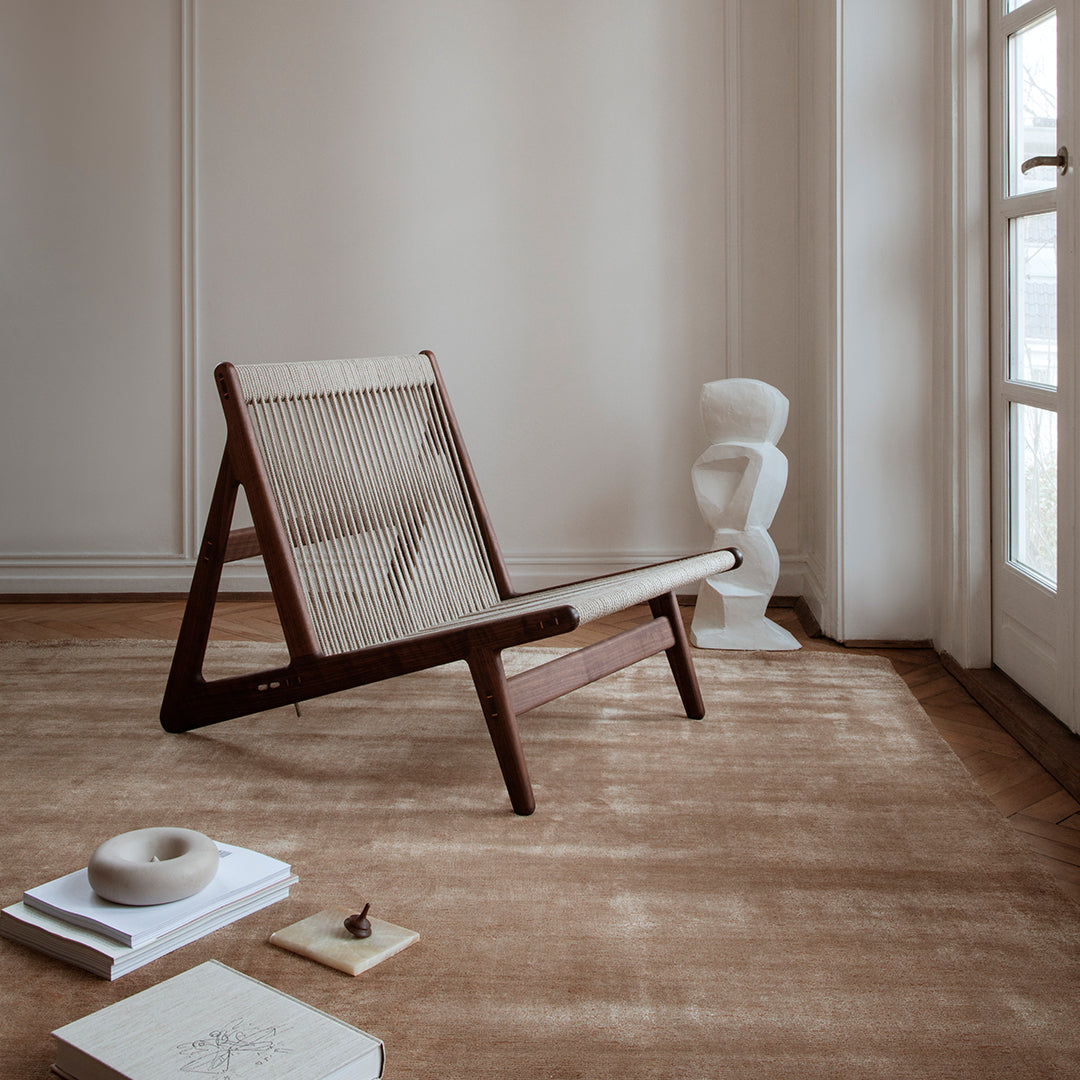 Modern wood lounge chair with beige fabric, set against a neutral-toned room with a textured brown rug.