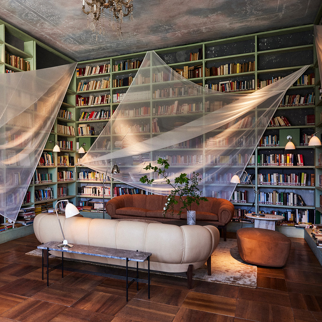 Cozy living room with green bookshelves, beige sofa, and sheer white fabric draping. Warm wood flooring adds elegance.