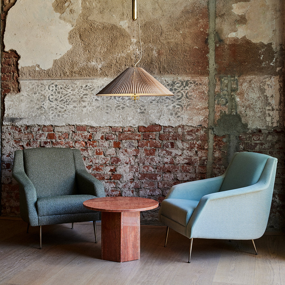 Pair of upholstered armchairs in soft blue and green hues, with a round wooden table against a textured brick wall.