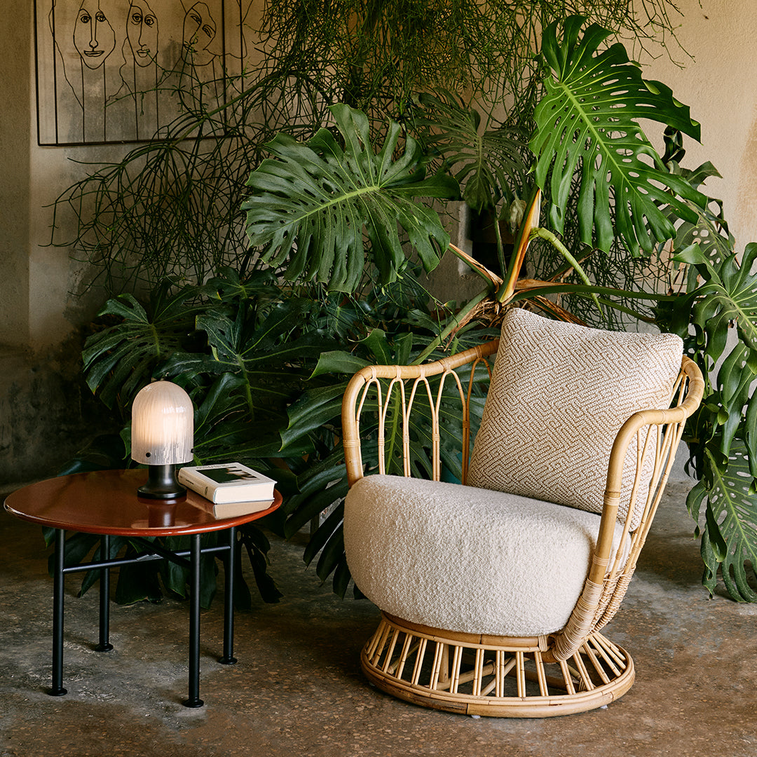 "Organic rattan chair with cream upholstery, set against lush greenery and a warm wooden side table."