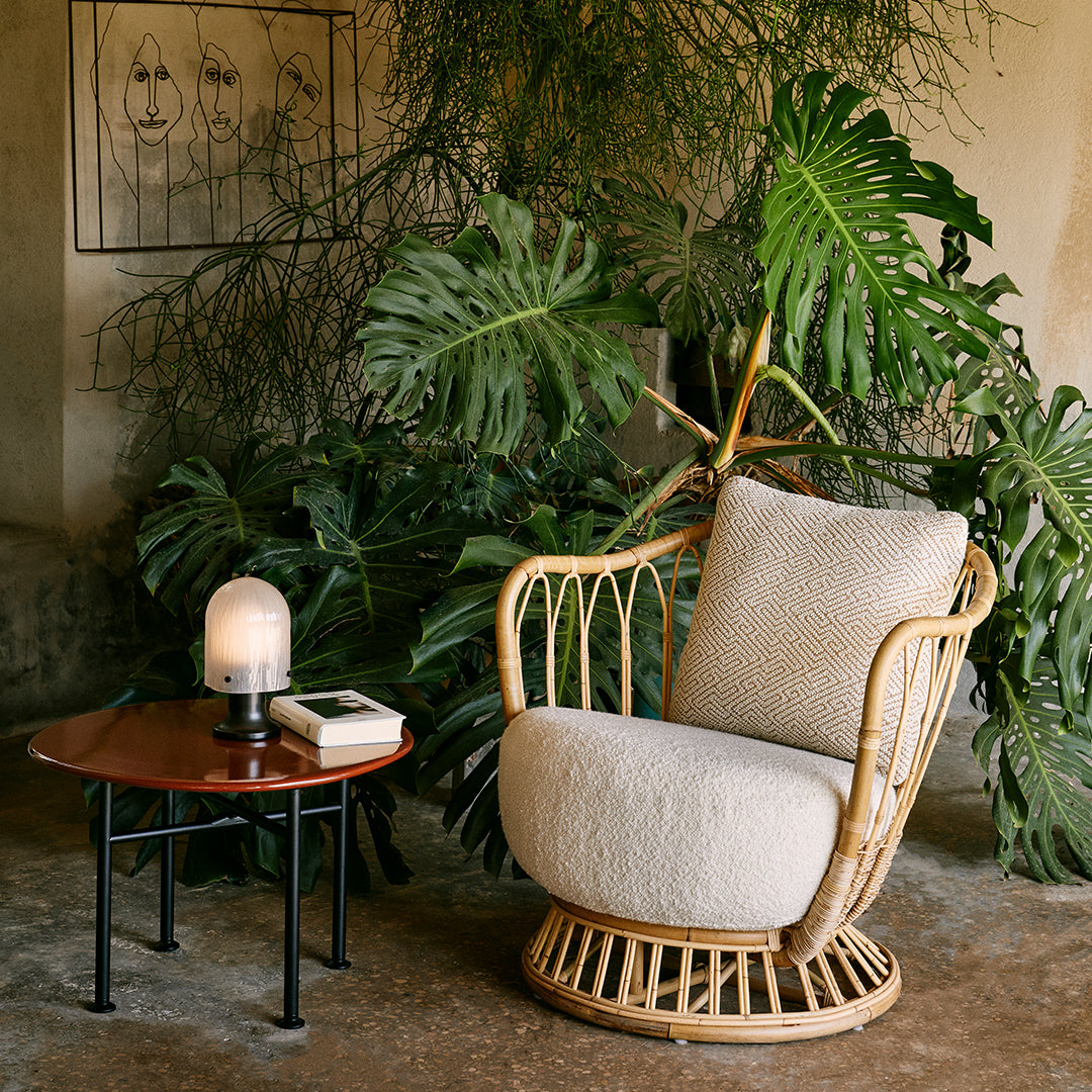 Neutral beige rocking chair with boho rattan design, set against lush greenery and a rich wooden side table.