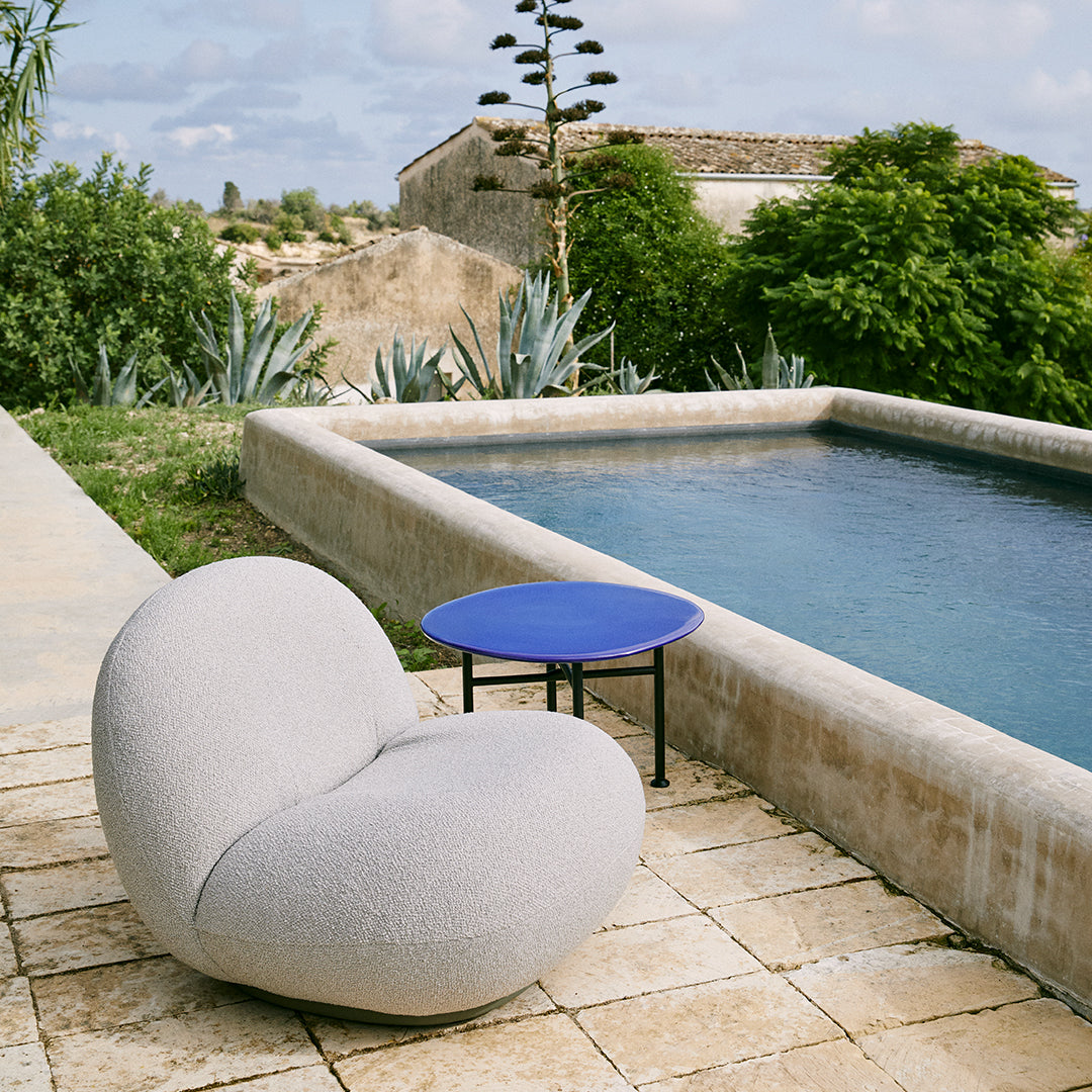 Gray curved chair with smooth texture and vibrant blue round table, set against a natural stone patio by a pool.