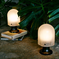 Two elegant white tabletop lamps with textured glass shades on black bases, surrounded by lush green foliage.