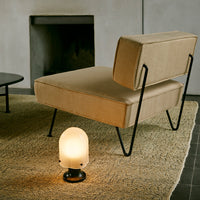 Beige upholstered accent chair with black metal legs, sitting on a textured tan rug beside a round table. Soft lighting.