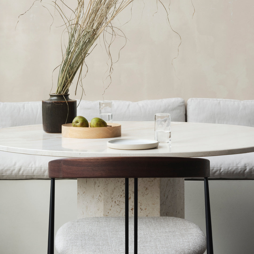 Round marble table with soft gray seating, black vase, wooden bowl, and green apples, creating a minimalist, elegant look.