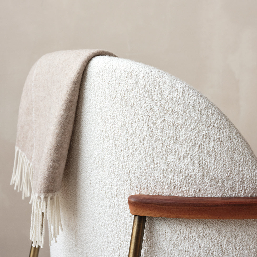 Beige textured throw blanket draped on a white boucle chair with elegant fringe detailing.