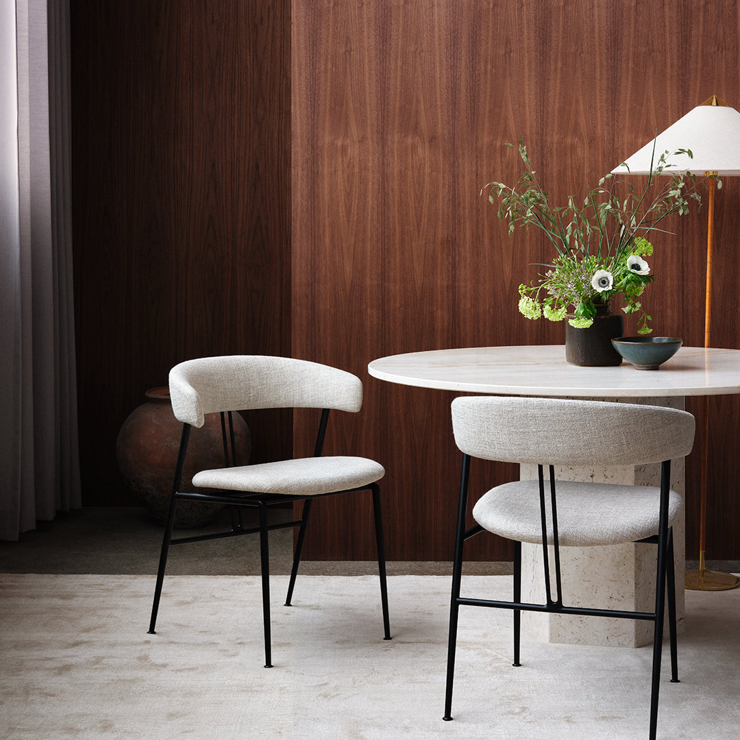 "Light gray upholstered chairs with black legs, round white table, and natural wood backdrop featuring subtle textures."