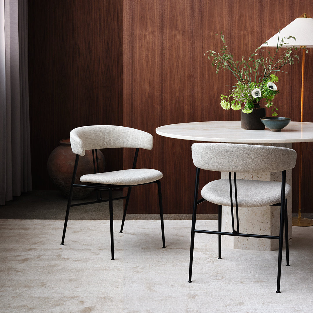 Neutral beige upholstered chairs with sleek black metal legs beside a light marble table, against a warm wood backdrop.
