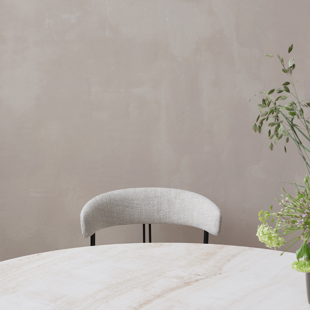 "Soft gray upholstered chair with a curved backrest, set against a muted beige wall and light marble table."