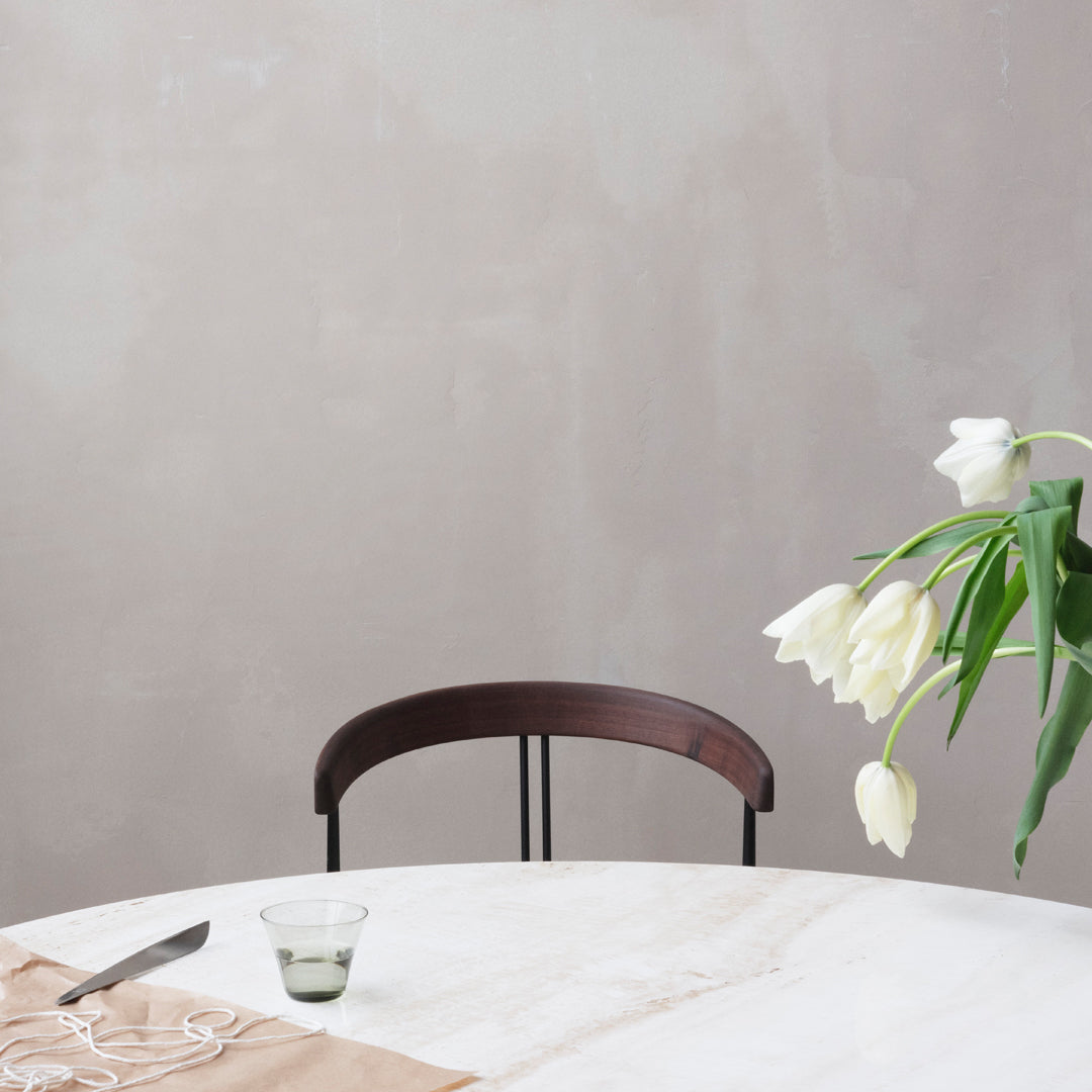 Elegant beige table with a soft marble pattern, dark wooden chair, and delicate white tulips in green vase.