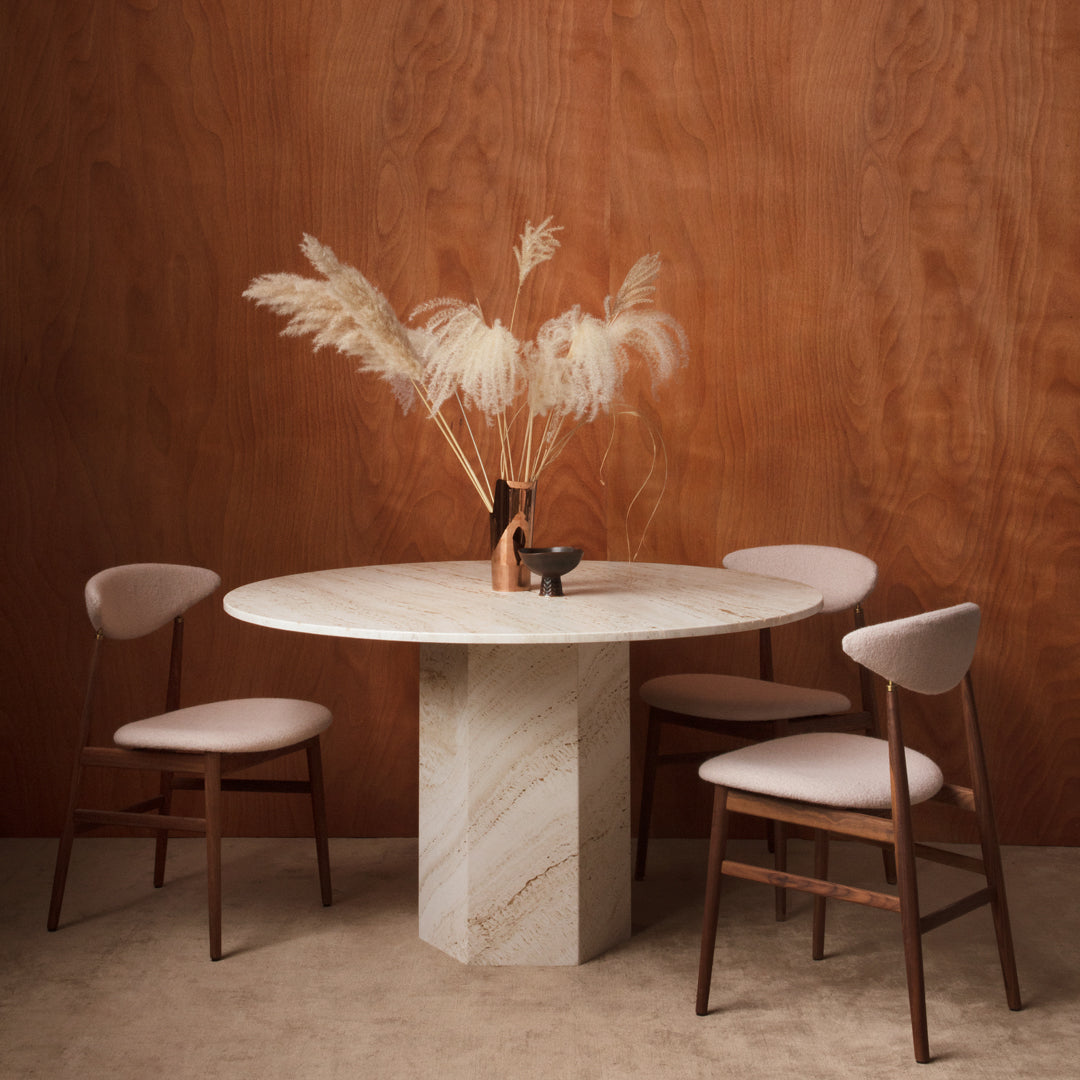 Round marble dining table with beige and cream tones, featuring soft pink upholstered chairs and a warm wooden backdrop.