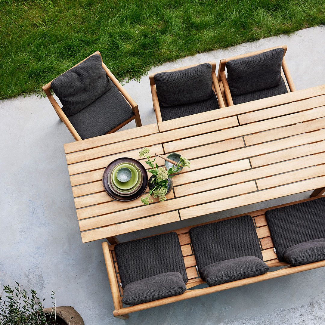 Wooden patio table with slatted design, surrounded by dark gray cushioned chairs and a green decorative bowl.