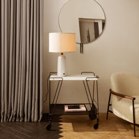 White tabletop on a black metal cart with a beige lamp; soft gray curtains and a round mirror in the background.
